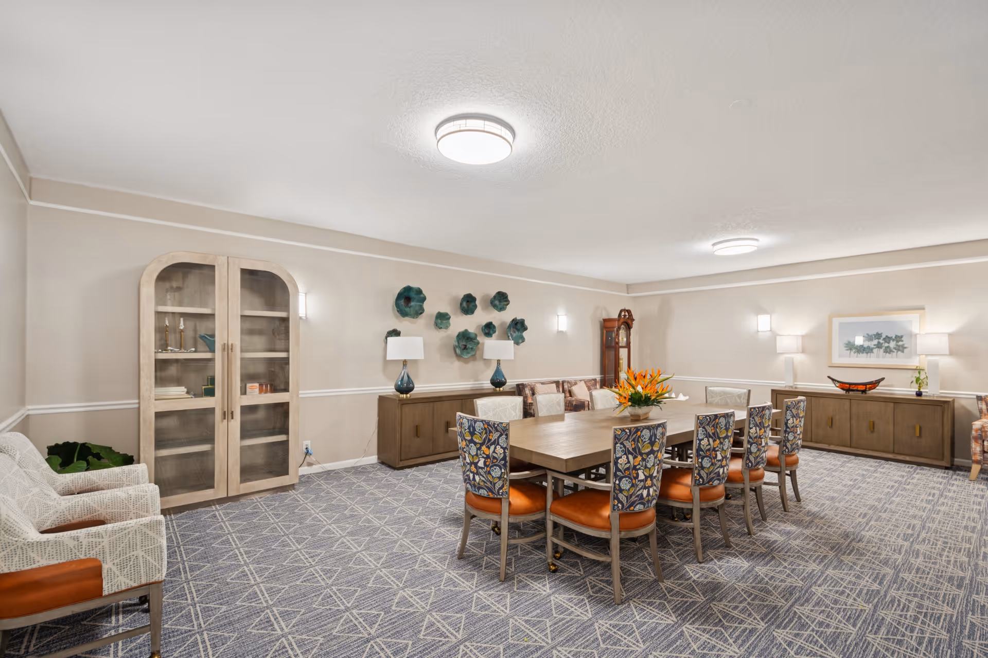 A spacious, well-lit dining room with a large rectangular wooden table surrounded by eight chairs with patterned backs and orange seats. The room features a patterned carpet, beige walls with white trim, two wooden cabinets, decorative wall art, table lamps, and a grandfather clock in the corner.