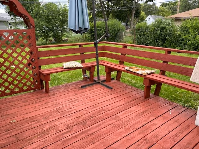 Outdoor wooden deck painted red with built-in corner bench seating and two cushions. A closed patio umbrella is positioned in the middle of the seating area. The deck overlooks a grassy backyard with trees and neighboring houses in the background.