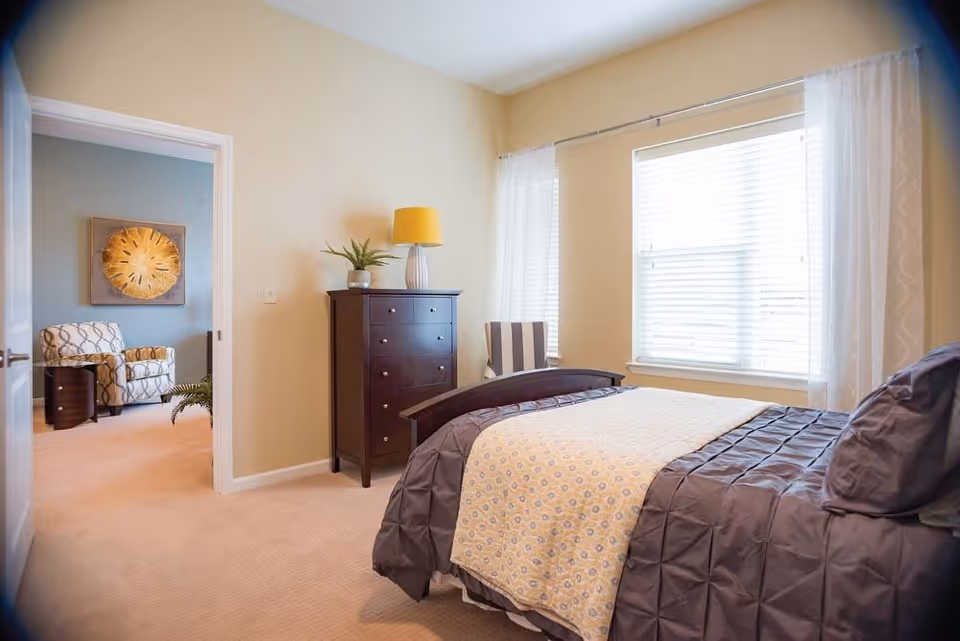 A bright bedroom with a bed covered in a gray comforter and a patterned yellow and white blanket. Next to the bed is a striped armchair by a window with white curtains. A dark wooden dresser with a yellow lamp and a small plant sits against the wall. An open door reveals an adjacent room with a patterned armchair, a small round table, and a decorative wall piece.