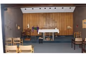 Small chapel interior with an altar draped in a cloth, wooden paneled wall, flags, and several chairs arranged facing the altar.