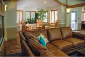 Interior view of a senior living facility common area with a large brown leather sectional sofa in the foreground, a round wooden table with chairs near windows with wooden blinds, and a potted plant. The room has wooden flooring and light-colored walls with wooden beams and trim.