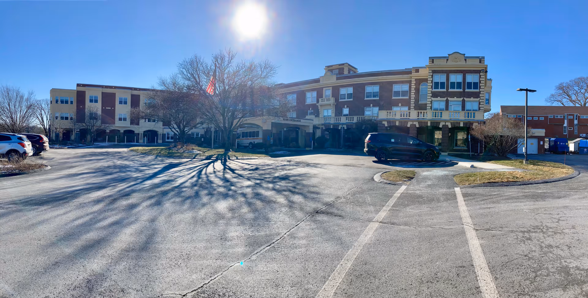 Exterior view of a multi-story brick building with a main entrance canopy, several parked cars, leafless trees casting long shadows on the pavement, and a clear blue sky with the sun shining brightly.