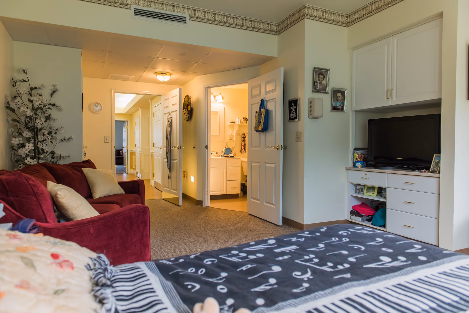 Interior view of a senior living facility room at Kingston Residence of Marion showing a bed with patterned blankets in the foreground, a red couch with pillows to the left, a TV on a white cabinet to the right, and an open door leading to a bathroom with a vanity and mirror. The hallway beyond the bathroom door is visible with additional doors and a clock on the wall.