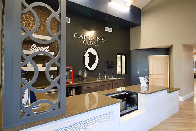 Interior view of a senior living facility area named Captain's Cove featuring a countertop with a built-in sink and beverage cooler, decorative shelving with the word 'Sweet', and a dark accent wall with the Captain's Cove logo and silhouette.