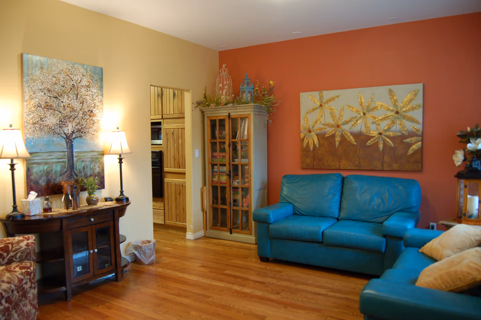 A cozy living room with wooden flooring, two teal leather sofas, a wooden cabinet with glass doors filled with books, and two paintings on the walls. There are two table lamps on a wooden side table next to a floral patterned armchair. The walls are painted in warm tones of beige and burnt orange.