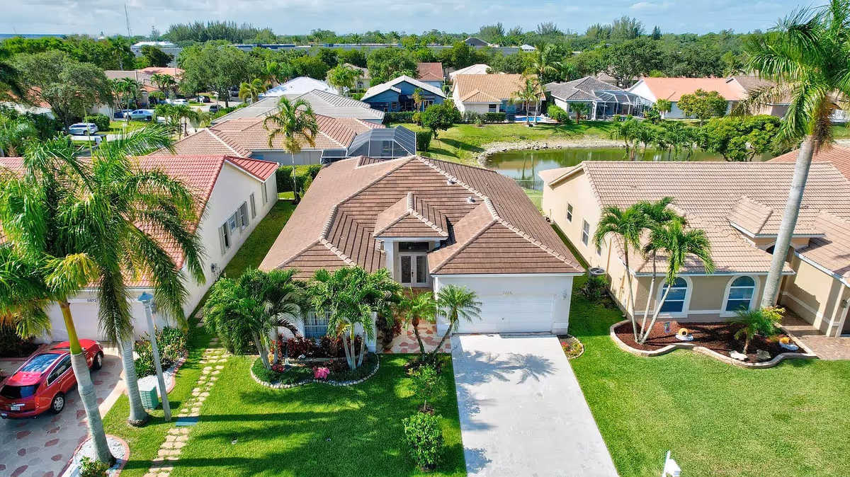 Aerial view of a residential neighborhood featuring single-story houses with tiled roofs, green lawns, palm trees, and a small pond in the background under a partly cloudy sky.