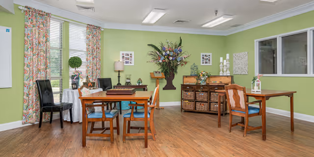 Bright communal dining/activity room with light green walls, wooden tables and chairs, floral decorations, and storage baskets.