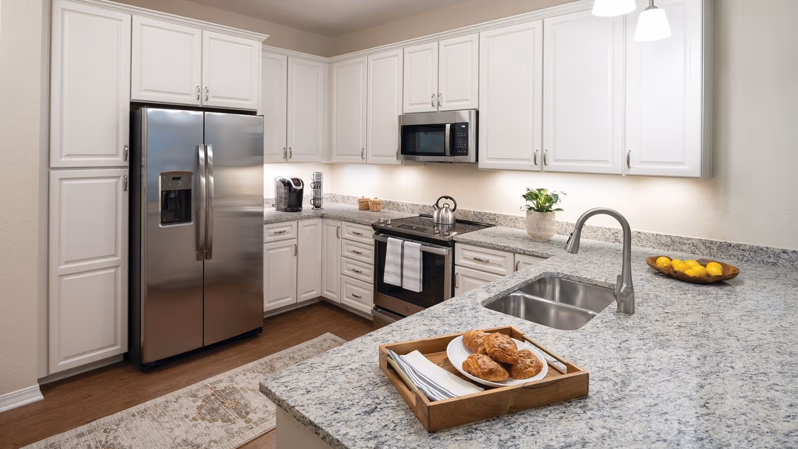 Modern kitchen with white cabinets, stainless steel refrigerator, oven, microwave, and a granite countertop with a sink. A wooden tray with pastries and napkins is placed on the counter, along with a bowl of lemons and a small potted plant.