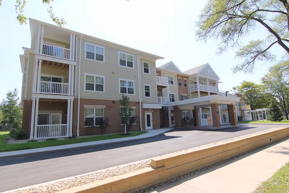 Exterior view of a three-story residential building with balconies and a covered entrance, surrounded by trees and a paved driveway, under a clear sky.
