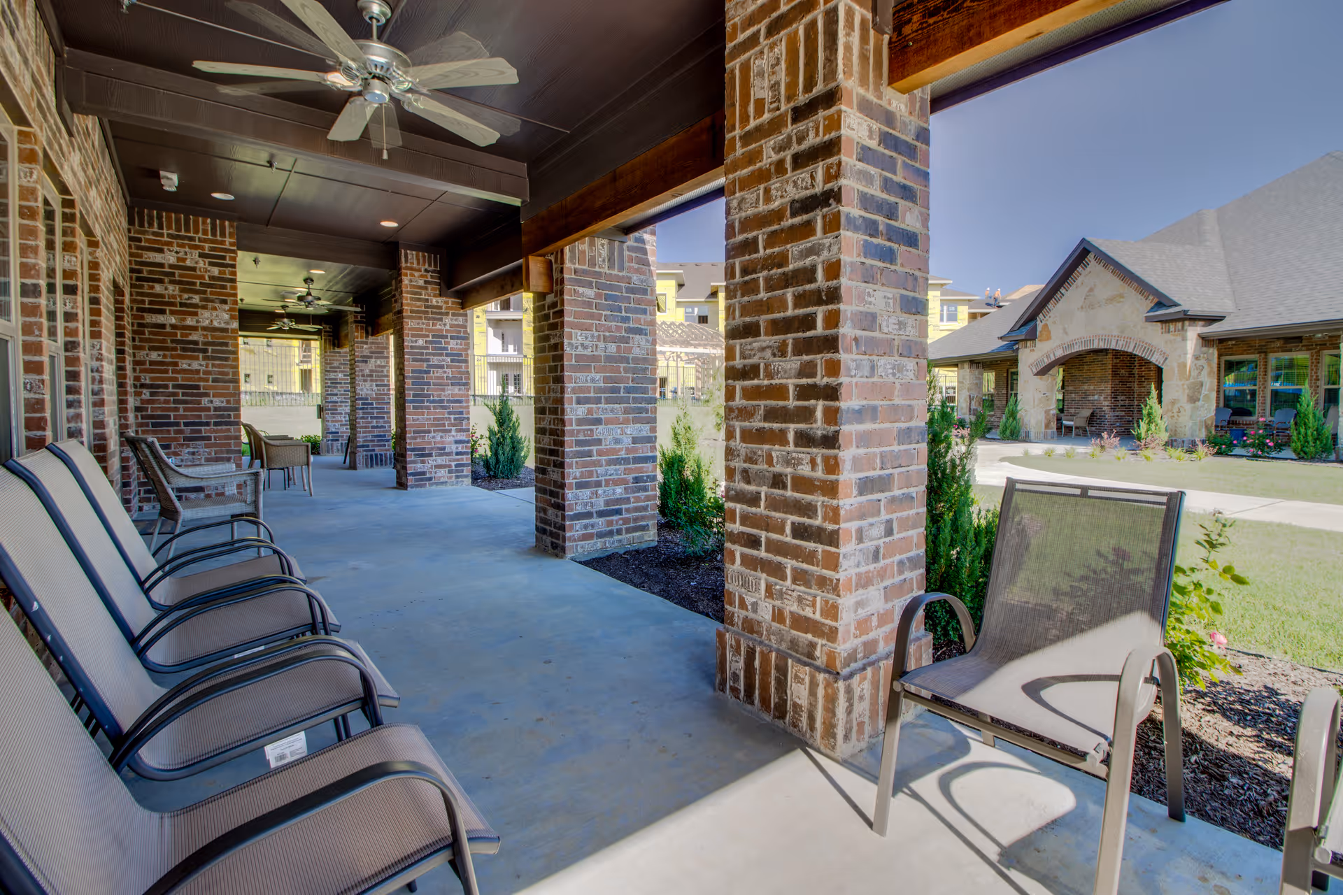 Covered outdoor patio area with brick columns and ceiling fans, featuring several chairs arranged along the walkway. In the background, there is a view of a landscaped garden and a building with a stone and brick exterior under a clear blue sky.