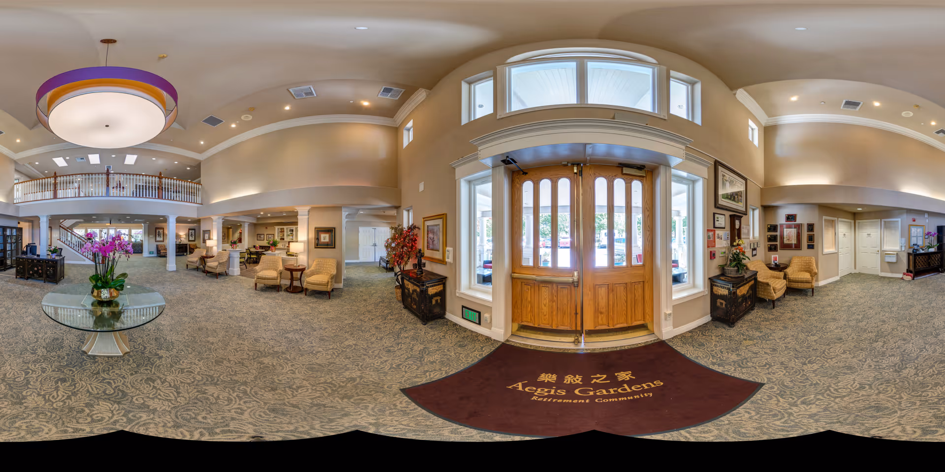 Spacious and well-lit lobby area of Aegis Gardens Fremont retirement community featuring a large circular chandelier, a round glass table with a flower arrangement, comfortable seating areas with armchairs, decorative plants, and large wooden double doors with windows letting in natural light.