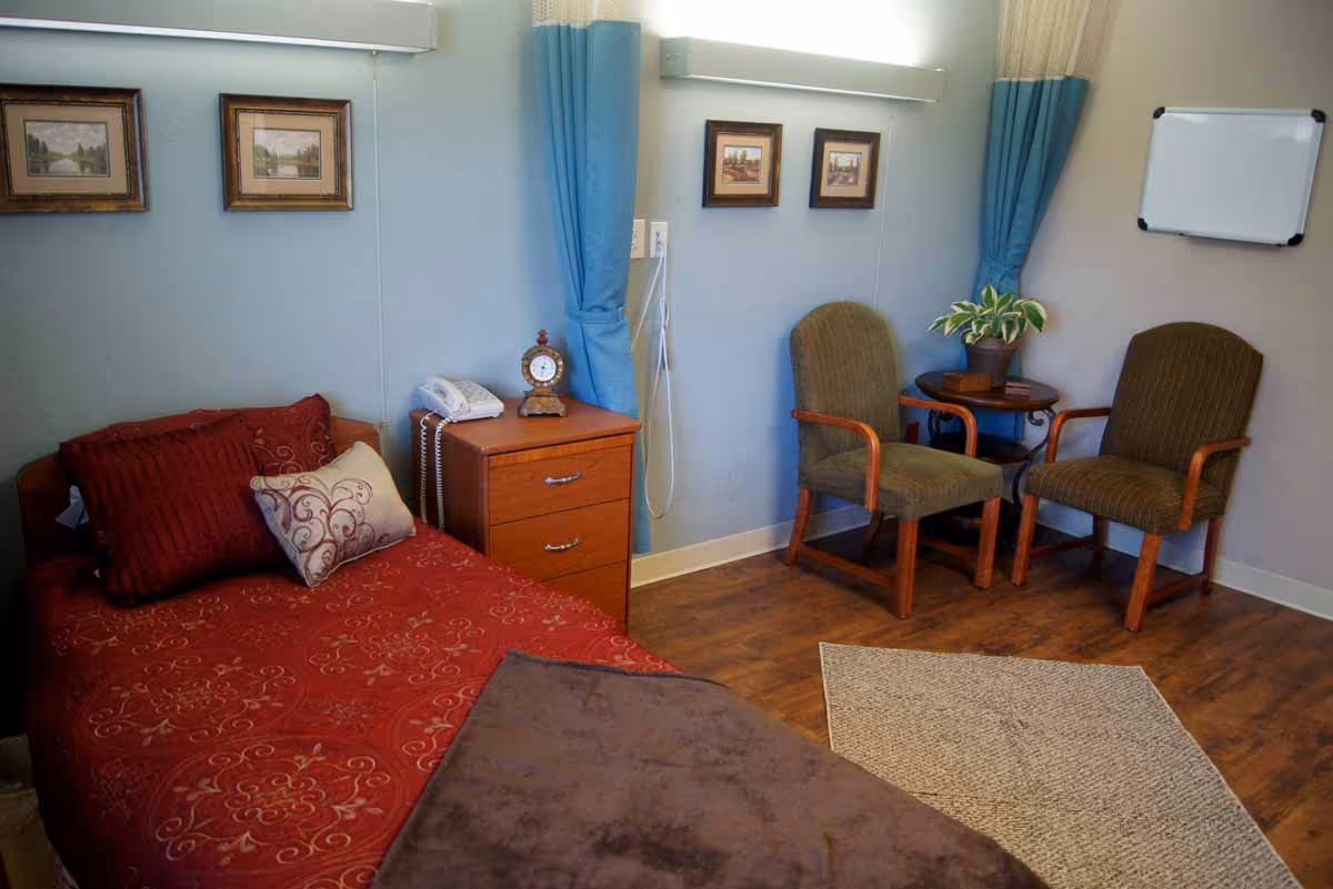 A cozy nursing facility room with a single bed covered in red bedding and decorative pillows. Next to the bed is a wooden nightstand with a telephone and a vintage-style clock. The room has two framed pictures on the wall above the bed and two more above a small round table with a potted plant. Two wooden armchairs with green cushions are placed around the table. A whiteboard is mounted on the wall, and the floor is wooden with a small area rug.