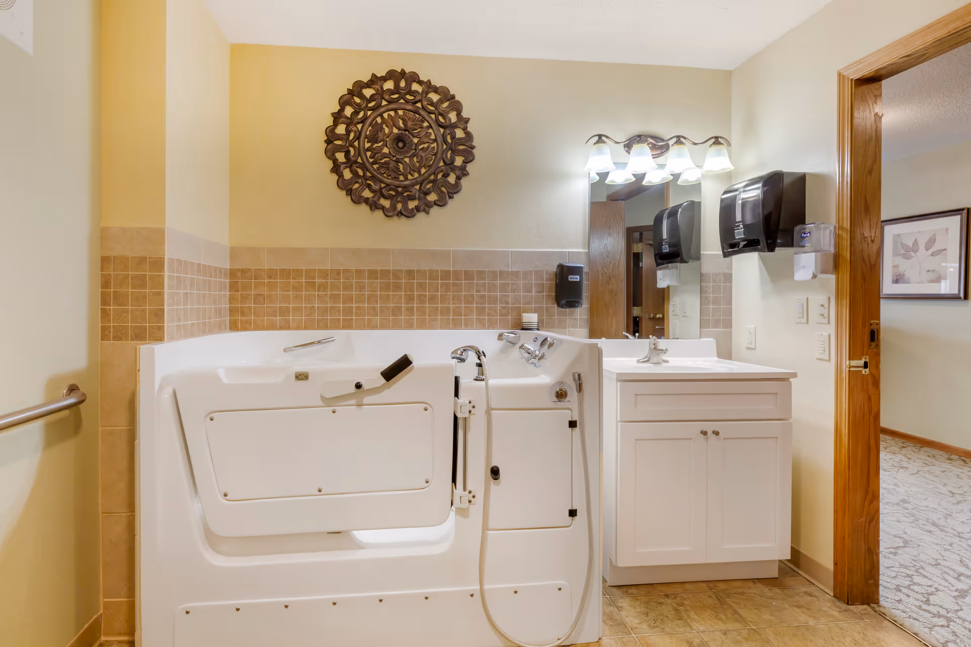 A bathroom featuring a walk-in bathtub with a door and safety handle, a white vanity with a sink and mirror above it, wall-mounted soap and paper towel dispensers, beige tiled walls, and a decorative circular wall hanging. The bathroom opens into a carpeted hallway with framed artwork on the wall.