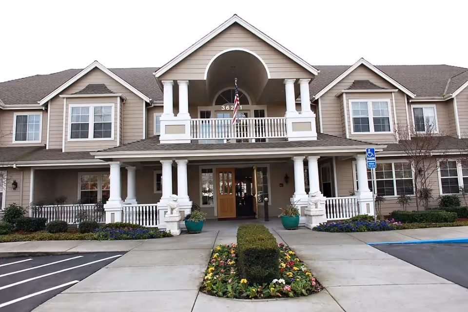 Front exterior view of a two-story senior living facility building with beige siding, white columns, and a covered entrance. There is an American flag hanging above the entrance, flower beds with colorful flowers, and a handicapped parking sign visible near the entrance.