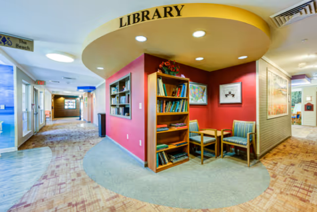 Interior view of a memory care community hallway featuring a small library nook with a wooden bookshelf filled with books, two striped chairs, and a small table. The library area is highlighted by a yellow curved ceiling with the word 'LIBRARY' written on it. The hallway has patterned carpet and leads to other rooms with visible exit signs and wall decorations.