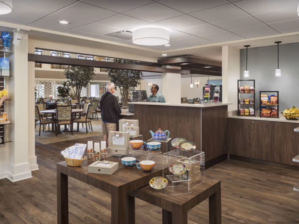 Interior view of a senior living facility's common area with a reception desk where a staff member is assisting an elderly man. In the foreground, there are wooden tables displaying decorative items such as bowls, a teapot, and picture frames. In the background, there are dining tables and chairs, plants, and shelves with snacks and bananas.