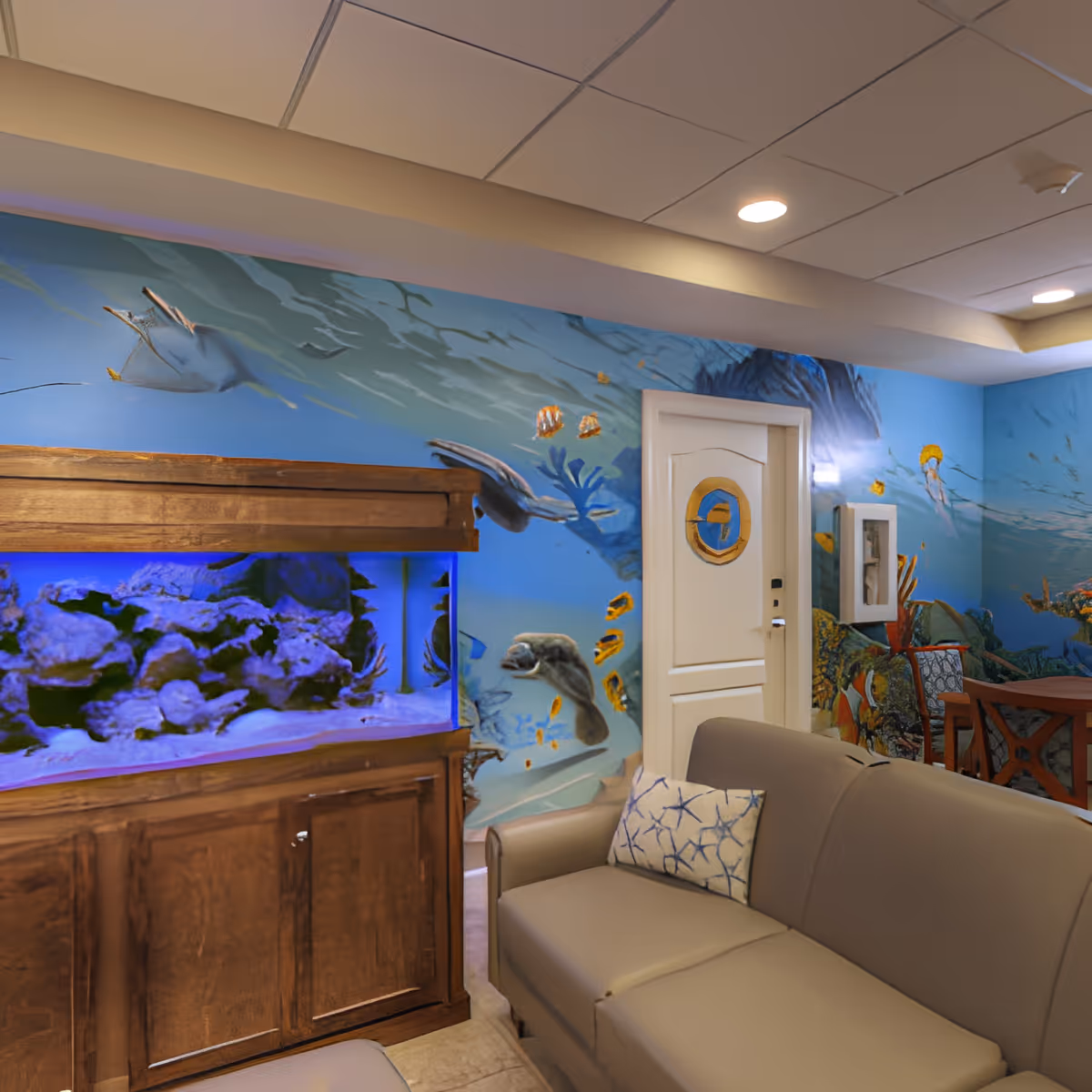 A cozy living room area with a beige sofa and a decorative pillow featuring a blue coral pattern. There is a large wooden aquarium filled with colorful coral and fish. The walls are painted with an underwater ocean mural depicting sea turtles, fish, and other marine life. A white door with a circular window is visible, along with a wooden table and chairs in the background.
