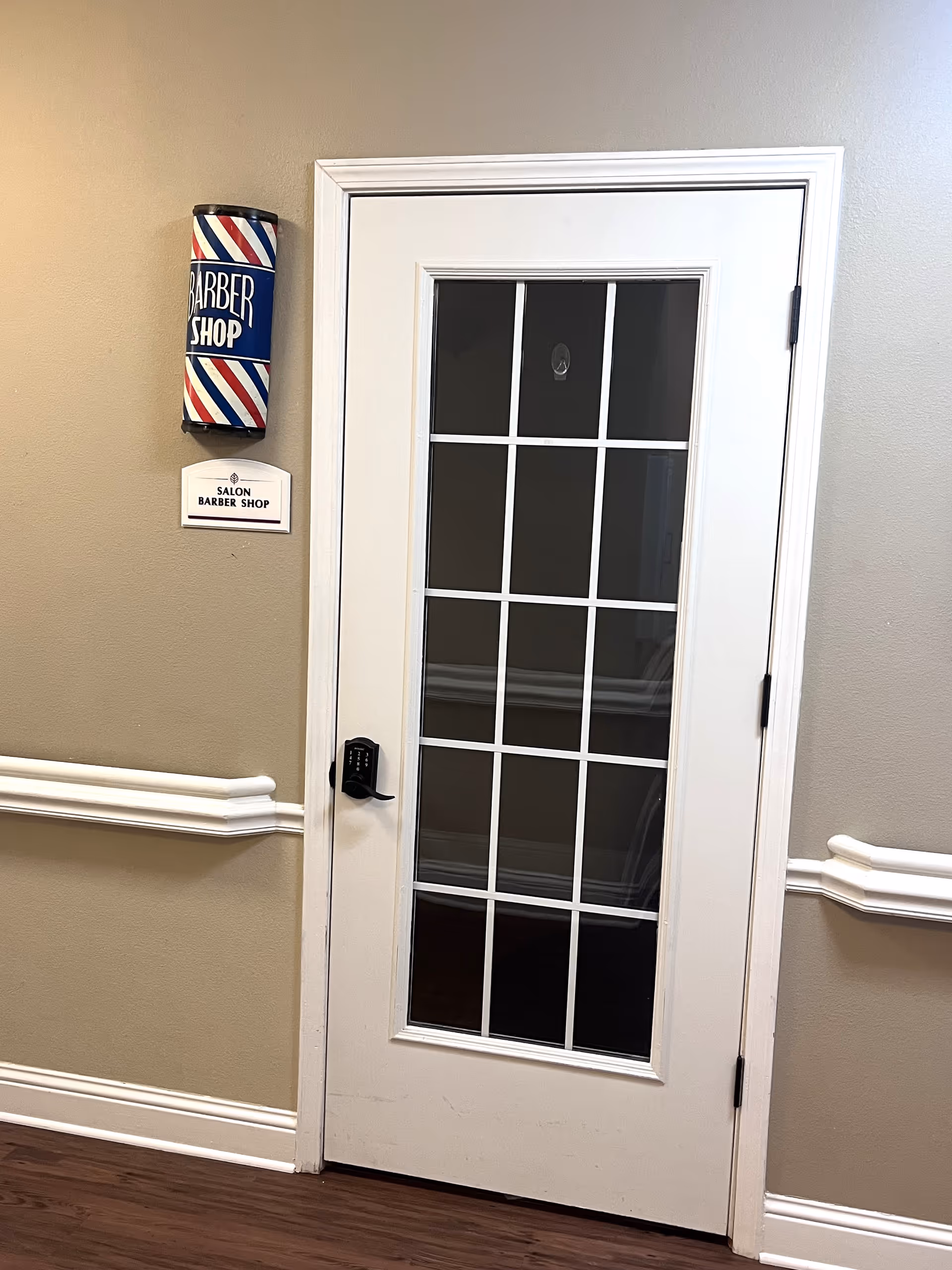 A white door with glass panes leading to a salon barber shop. To the left of the door, there is a traditional barber pole sign with red, white, and blue stripes and a small sign below it that reads 'SALON BARBER SHOP'. The wall is painted beige with white trim and molding, and the floor is dark wood.
