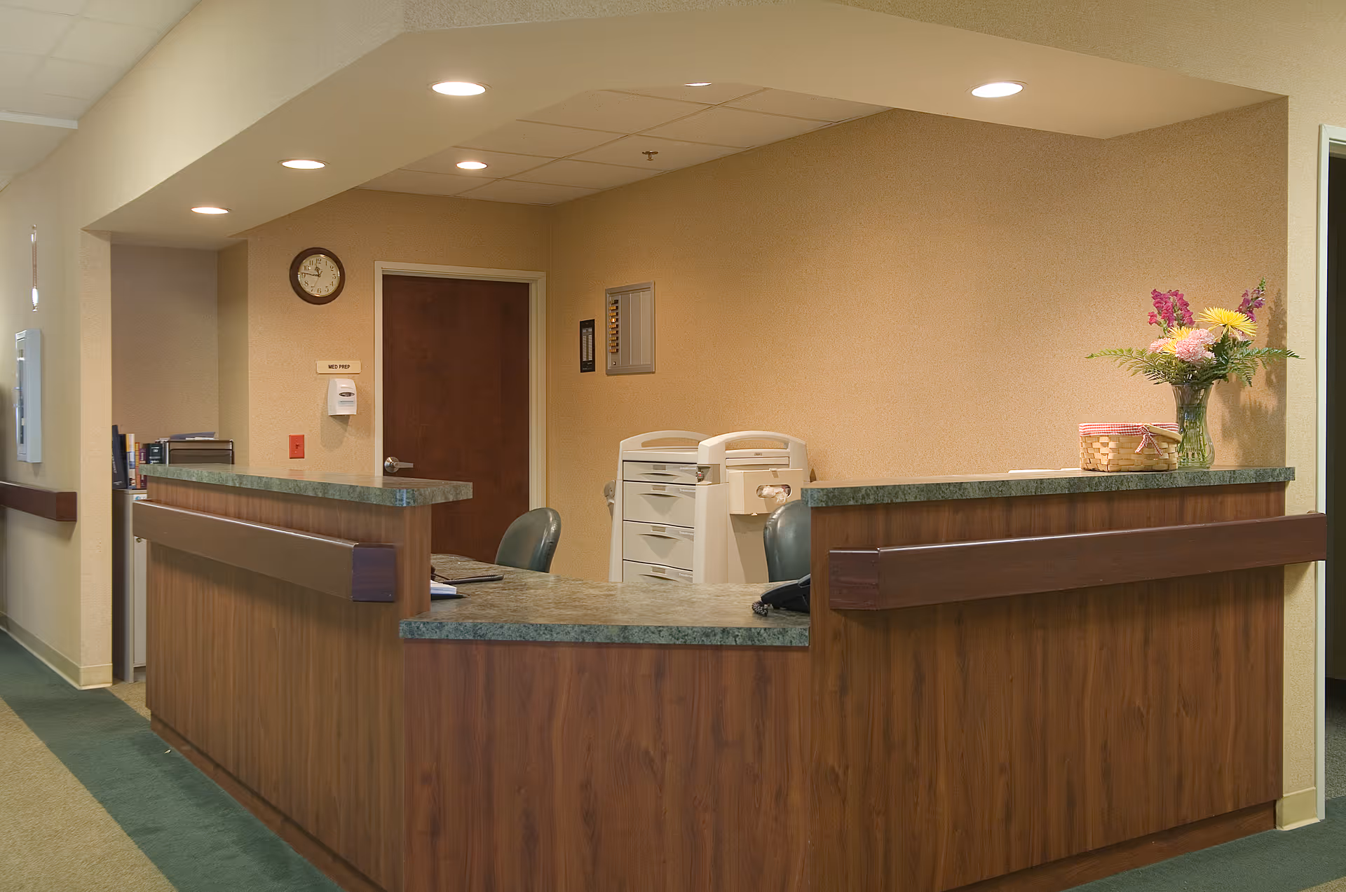 Reception desk area in a senior living facility with a wooden counter, chairs, office equipment, and a vase of flowers.