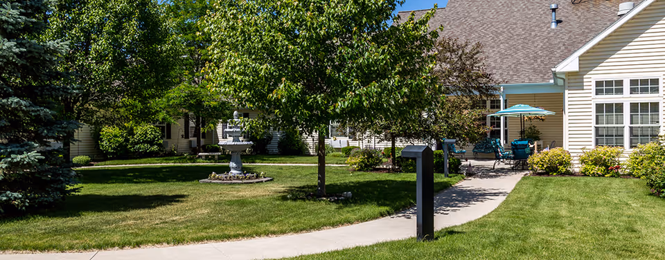 A well-maintained outdoor garden area with green grass, trees, and shrubs surrounding a concrete pathway. There is a decorative fountain in the center of the garden and patio furniture with an umbrella near the building entrance. The building has beige siding and multiple windows.
