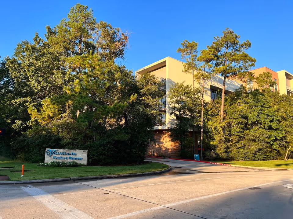 Exterior view of the Village at Woodlands Waterway building partially obscured by trees with a stone entrance sign by the road.