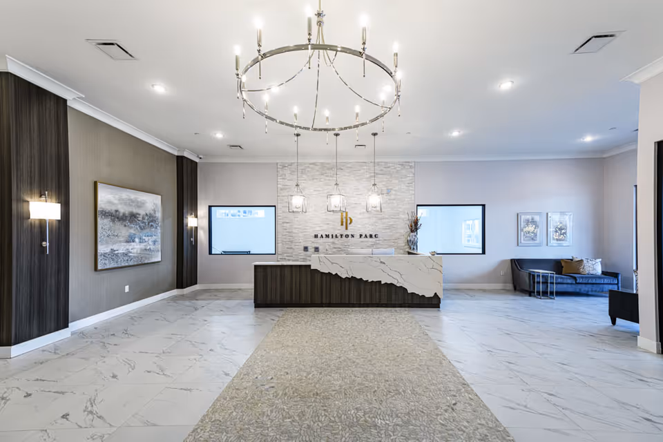 Spacious and modern reception area of Hamilton Parc featuring a large marble and wood front desk, a circular chandelier with multiple lights hanging from the ceiling, two windows behind the desk, a gray sofa with cushions on the right side, wall-mounted lights, and decorative artwork on the walls. The floor is tiled with a combination of marble and textured stone.