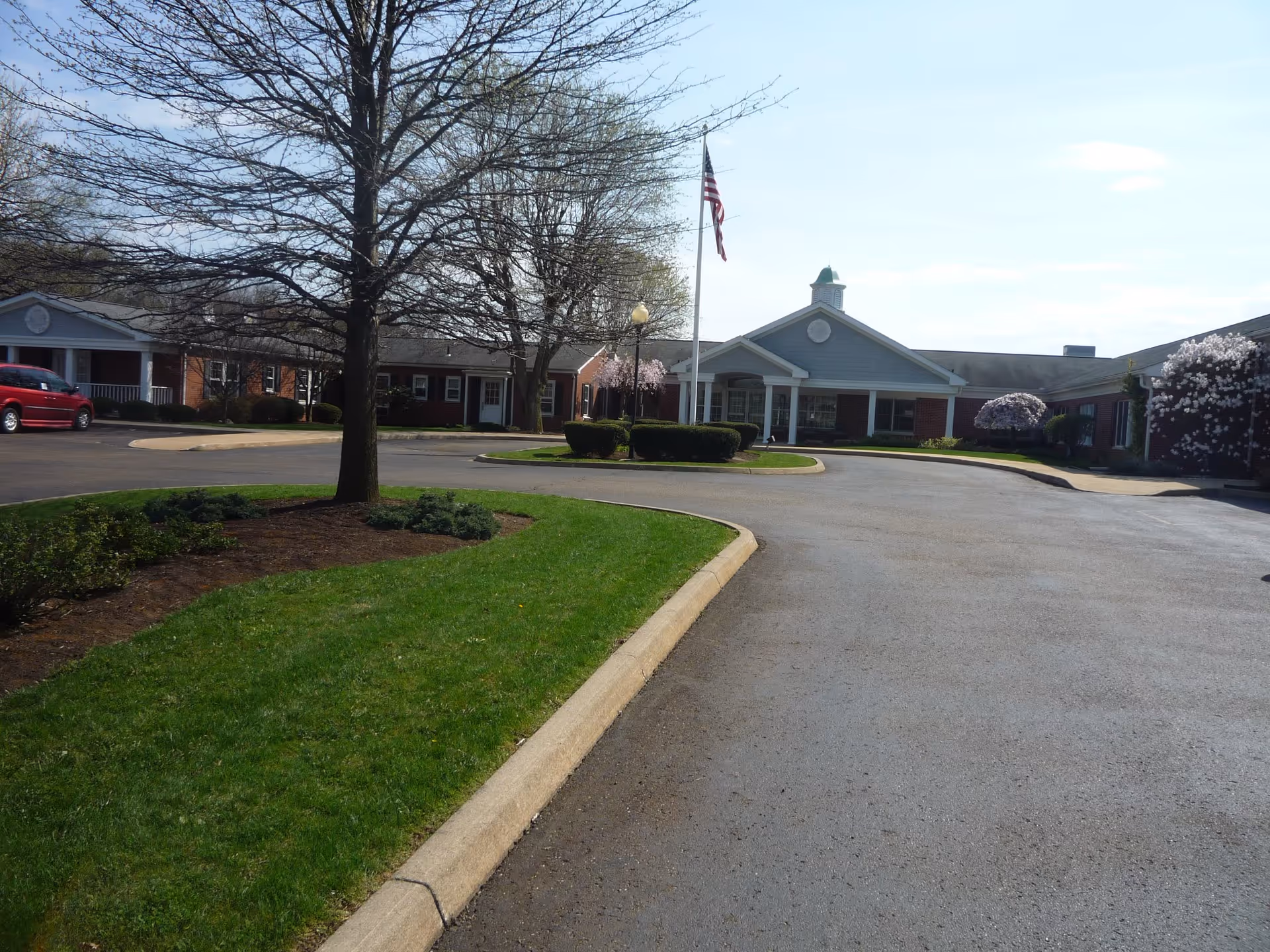 View of the front exterior of Windsor Medical Center showing a circular driveway with a flagpole in the center, surrounded by trimmed bushes and trees with some blooming flowers. The building has a brick facade with white columns and a cupola on the roof.