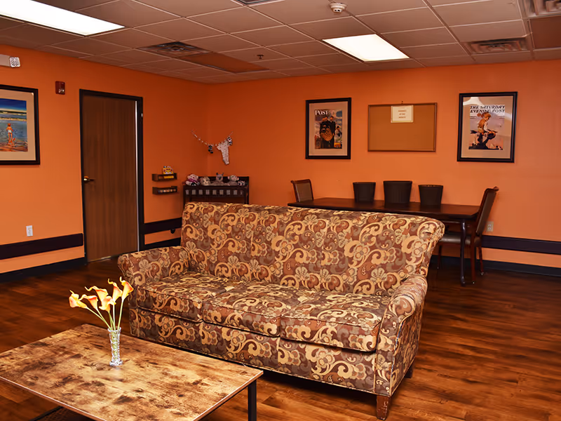 A cozy living room with an ornate patterned sofa in the center, a wooden coffee table with a small vase of flowers, orange walls adorned with framed pictures, a wooden door, and a table with chairs in the background.