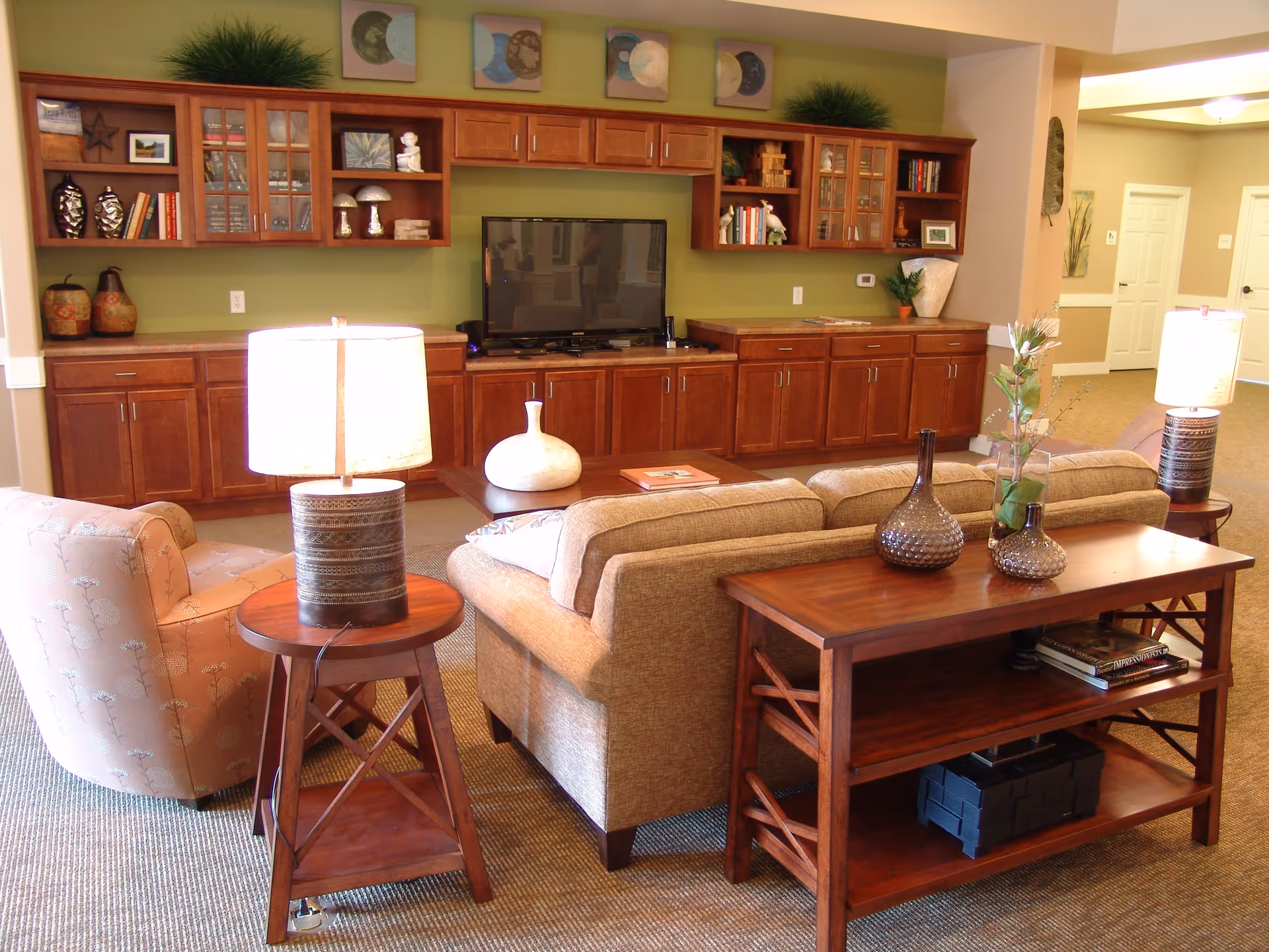 A cozy living room area with a beige sofa and a patterned armchair. There are two wooden side tables with lamps on either side of the sofa. Behind the sofa is a wooden console table with decorative vases and books. The back wall features built-in wooden cabinets with shelves holding books, decorative items, and a flat-screen TV in the center. The walls are painted green and beige, and the room has carpeted flooring.