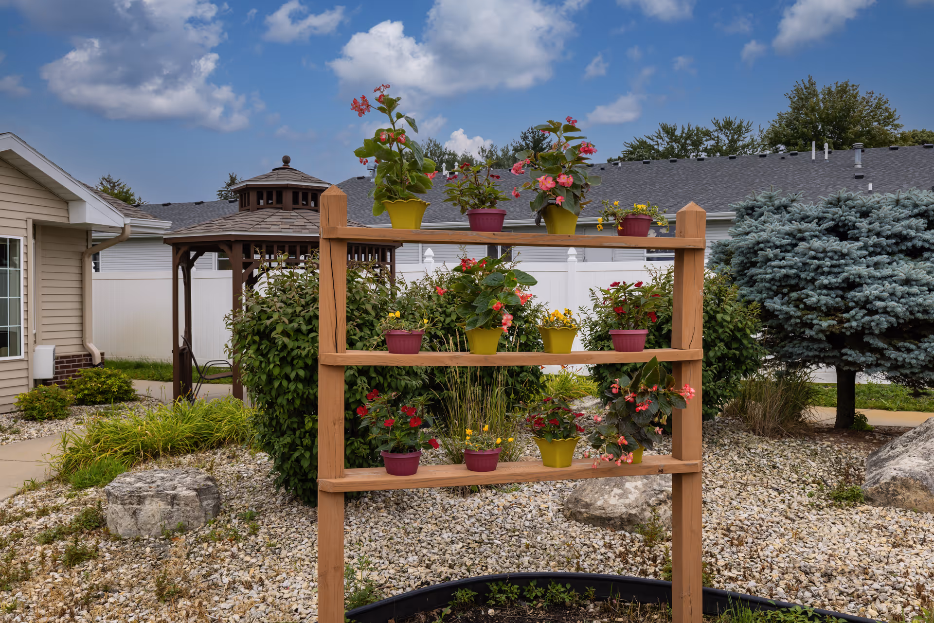 Outdoor garden area at Prairie Meadows Senior Living featuring a wooden plant stand with multiple colorful potted flowers. In the background, there is a gazebo, bushes, a tree, and residential buildings under a partly cloudy sky.