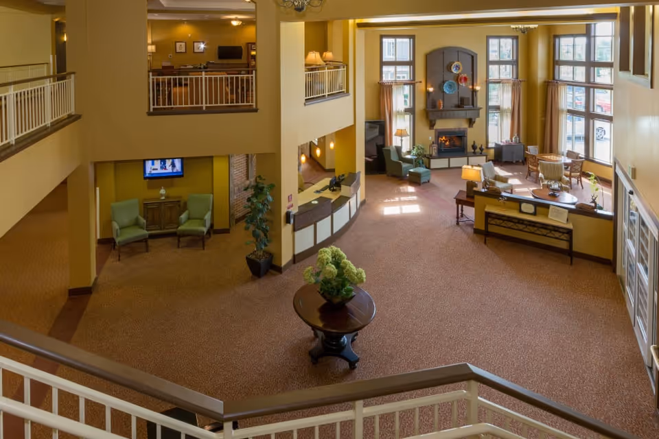 Spacious senior living facility lobby with high ceilings, large windows letting in natural light, a fireplace with decorative plates above it, comfortable seating areas with armchairs and tables, a reception desk, and a round table with a flower arrangement in the center.
