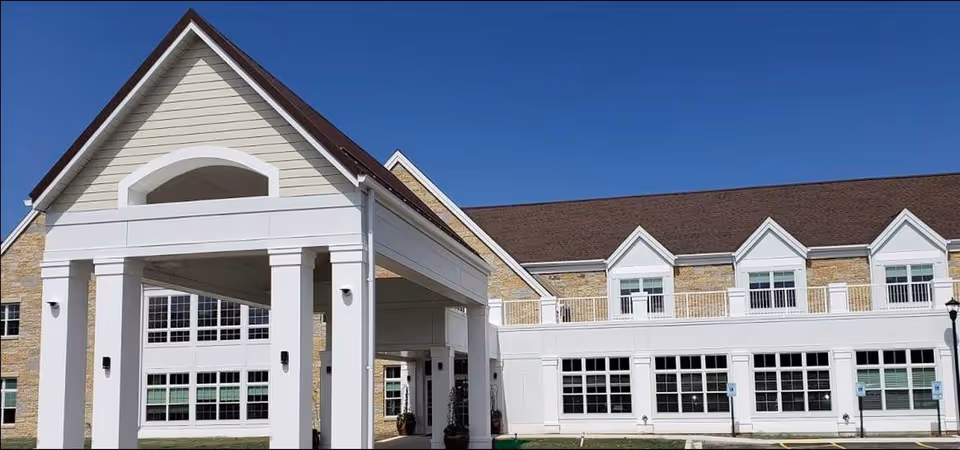 Exterior view of Silverado North Shore Memory Care Community building under a clear blue sky, featuring a covered entrance with white pillars and a beige and stone facade with multiple windows.