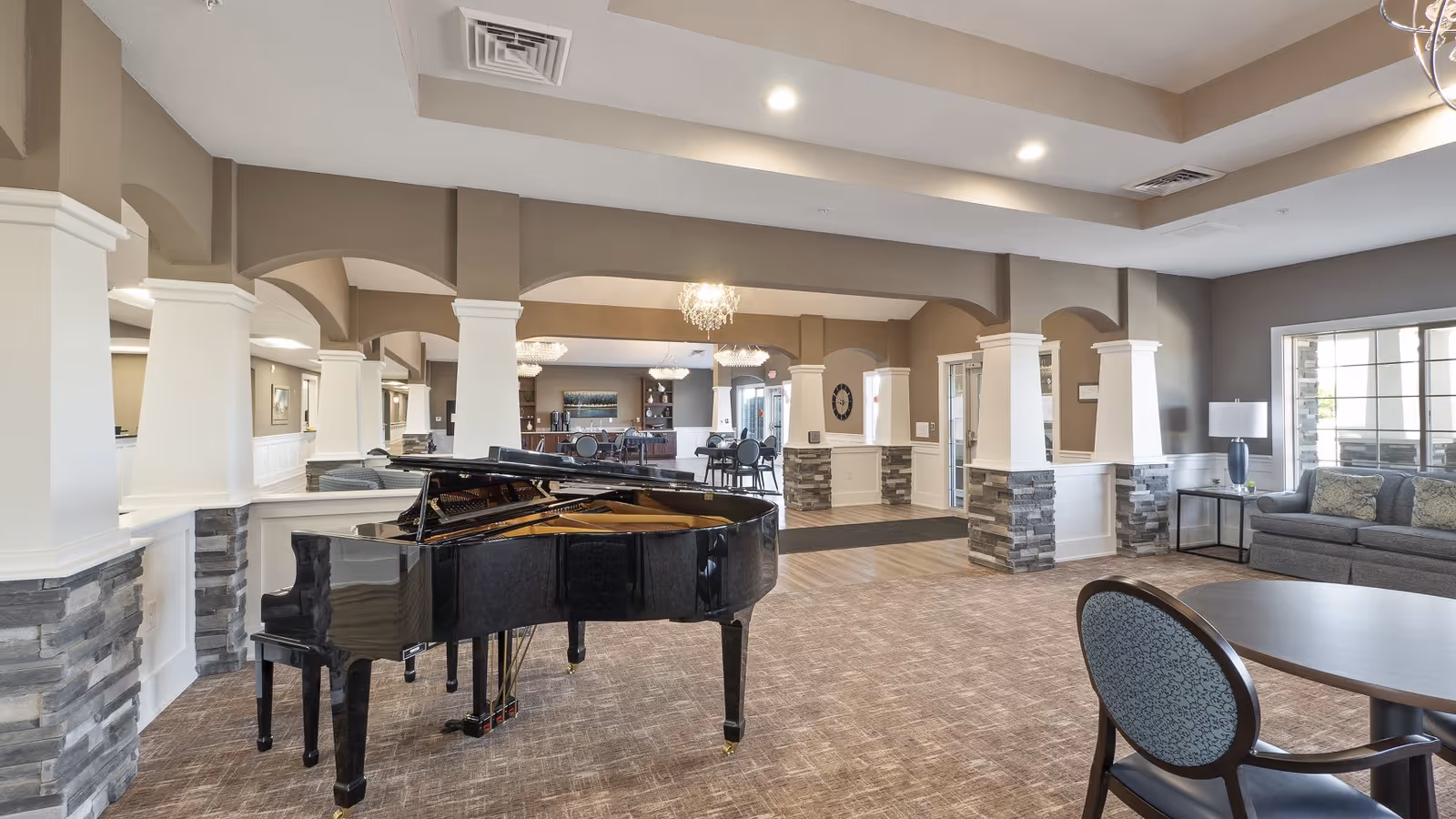 Spacious and well-lit common area in an assisted living facility featuring a black grand piano in the foreground, multiple seating areas with chairs and sofas, decorative columns with stone accents, and large windows allowing natural light to fill the room.