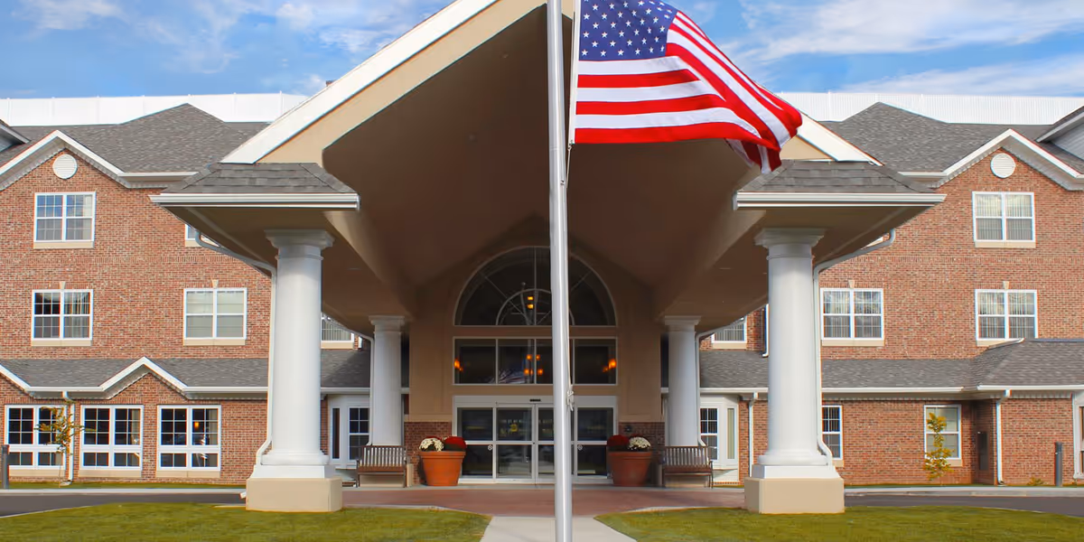 Front entrance of a brick retirement community with a covered portico supported by white columns and an American flag on a pole.