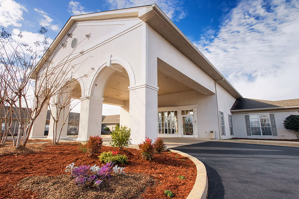 Entrance of a single-story care facility with a covered drive-through portico, arched columns, landscaped flowerbed, and clear sky.