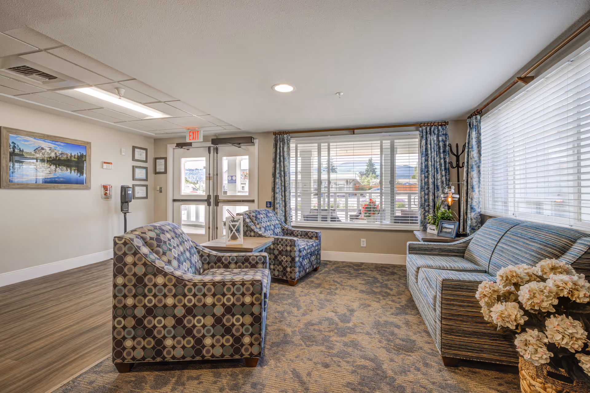 Lobby seating area with patterned armchairs, a striped sofa, a coffee table, large windows, and decorative flowers.