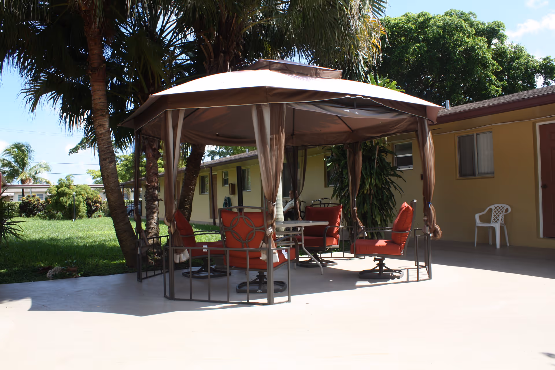 Outdoor patio area at Avalon Park Retirement Residence featuring a gazebo with a beige canopy and four red cushioned chairs around a small table. The patio is surrounded by green grass, palm trees, and a yellow building with windows and a door.
