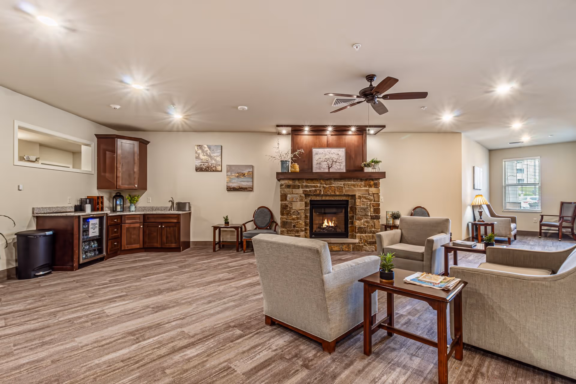A cozy assisted living common area featuring a stone fireplace with a wooden mantle, surrounded by beige upholstered armchairs and wooden side tables with small plants and magazines. The room has wood flooring, recessed ceiling lights, a ceiling fan, and a small kitchenette with dark wood cabinets and a mini fridge. There are framed pictures on the walls and a window letting in natural light.