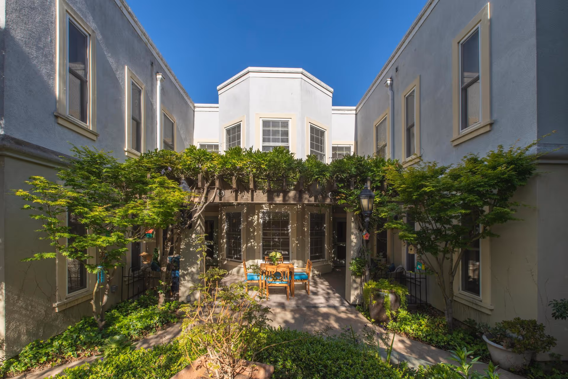 Outdoor courtyard area at Lakeside Park facility with a wooden pergola covered in greenery, surrounded by two-story building walls with multiple windows. There is a table with chairs under the pergola and various plants and trees around the courtyard.