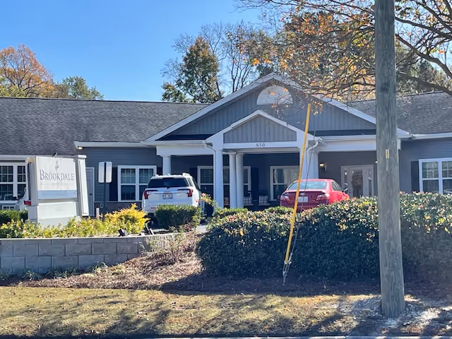 Exterior view of Brookdale Smithfield senior living facility showing a single-story building with a covered entrance, two parked cars, bushes, and trees with autumn foliage under a clear blue sky.