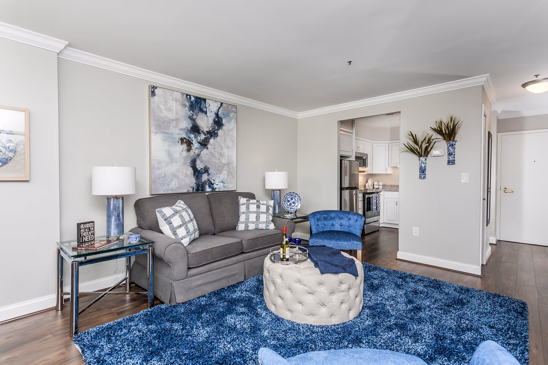 A modern senior living room with a gray sofa adorned with patterned pillows, a blue armchair, and a round tufted ottoman on a plush blue rug. The room features light gray walls with abstract artwork above the sofa, two blue and white vases with plants mounted on the wall, and a glimpse into a white kitchen with stainless steel appliances. There are two table lamps on glass side tables and wooden flooring throughout.
