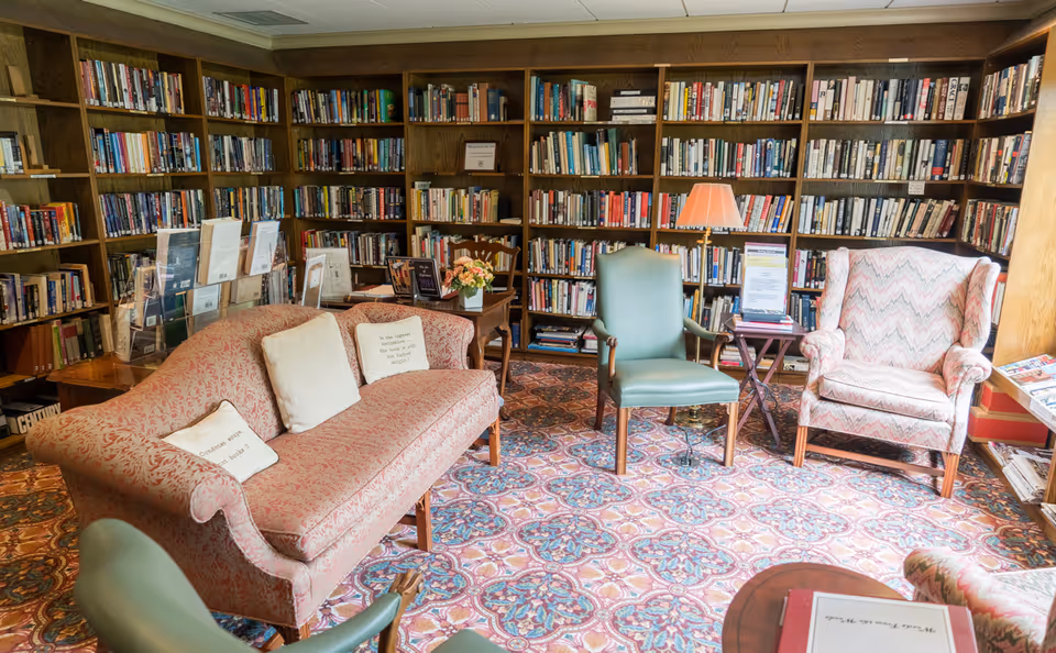 A cozy library room with wooden bookshelves filled with books lining the walls. There is a patterned carpet on the floor, a pink upholstered sofa with decorative pillows, two green armchairs, and a patterned wingback chair. A small wooden table with a lamp and some papers is also visible.