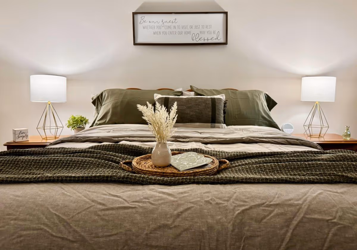 A neatly made bed with green and beige bedding, two pillows, and a decorative tray holding a small vase with dried flowers and a book. On either side of the bed are wooden nightstands with modern lamps, a small plant, and decorative items. Above the bed is a framed sign with an inspirational quote.