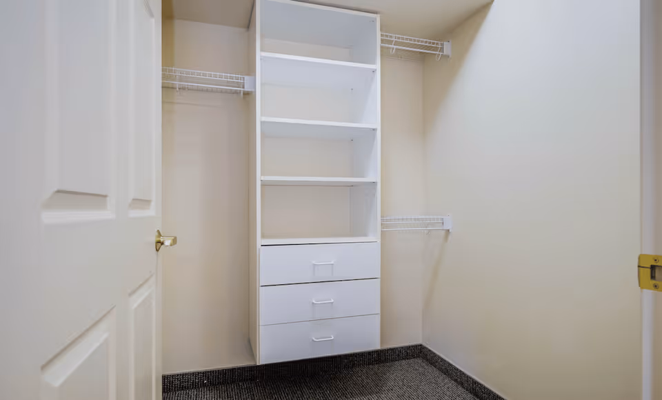 Empty walk-in closet with beige walls, a white shelving unit with three drawers, and wire racks for hanging clothes on the sides.