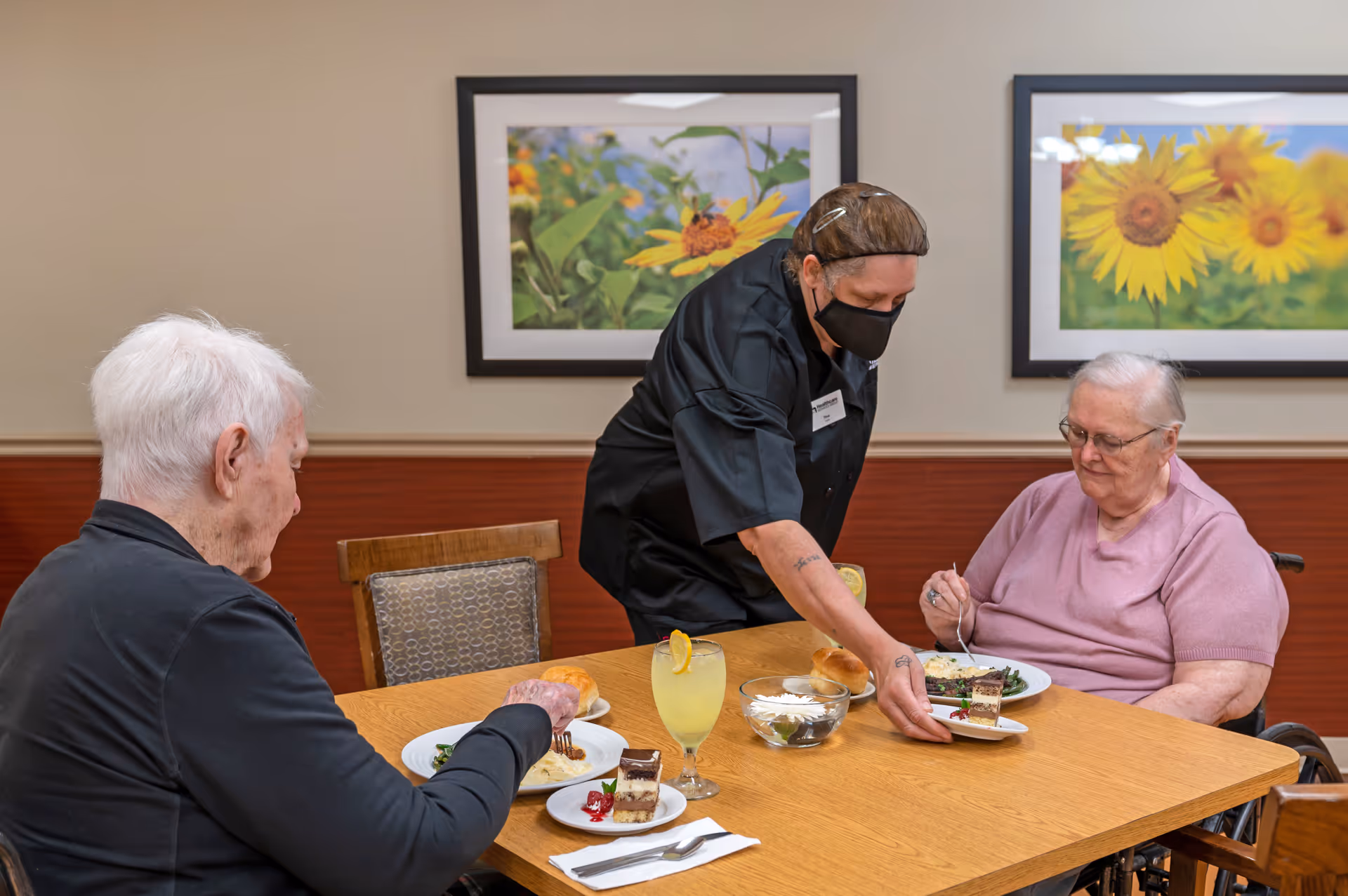 A dining room scene at Lakeland Rehabilitation & Health Care Center where a staff member wearing a black mask and uniform serves dessert to an elderly woman in a wheelchair. Another elderly person is seated at the table with plates of food, a glass of lemonade, and a bread roll. The room has framed pictures of sunflowers on the wall.