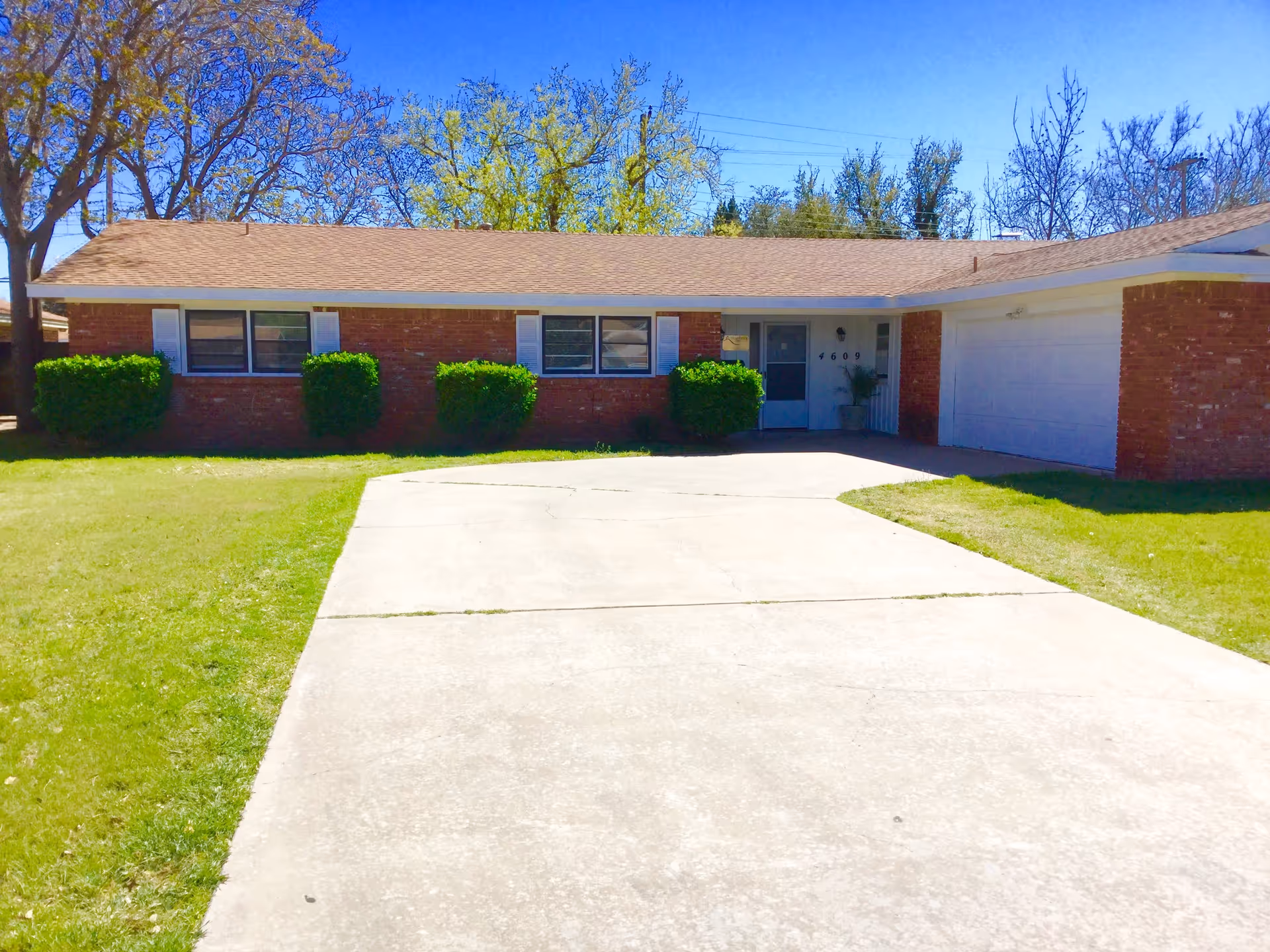 Front view of a single-story brick house with a concrete driveway, attached garage, lawn, and shrubs.