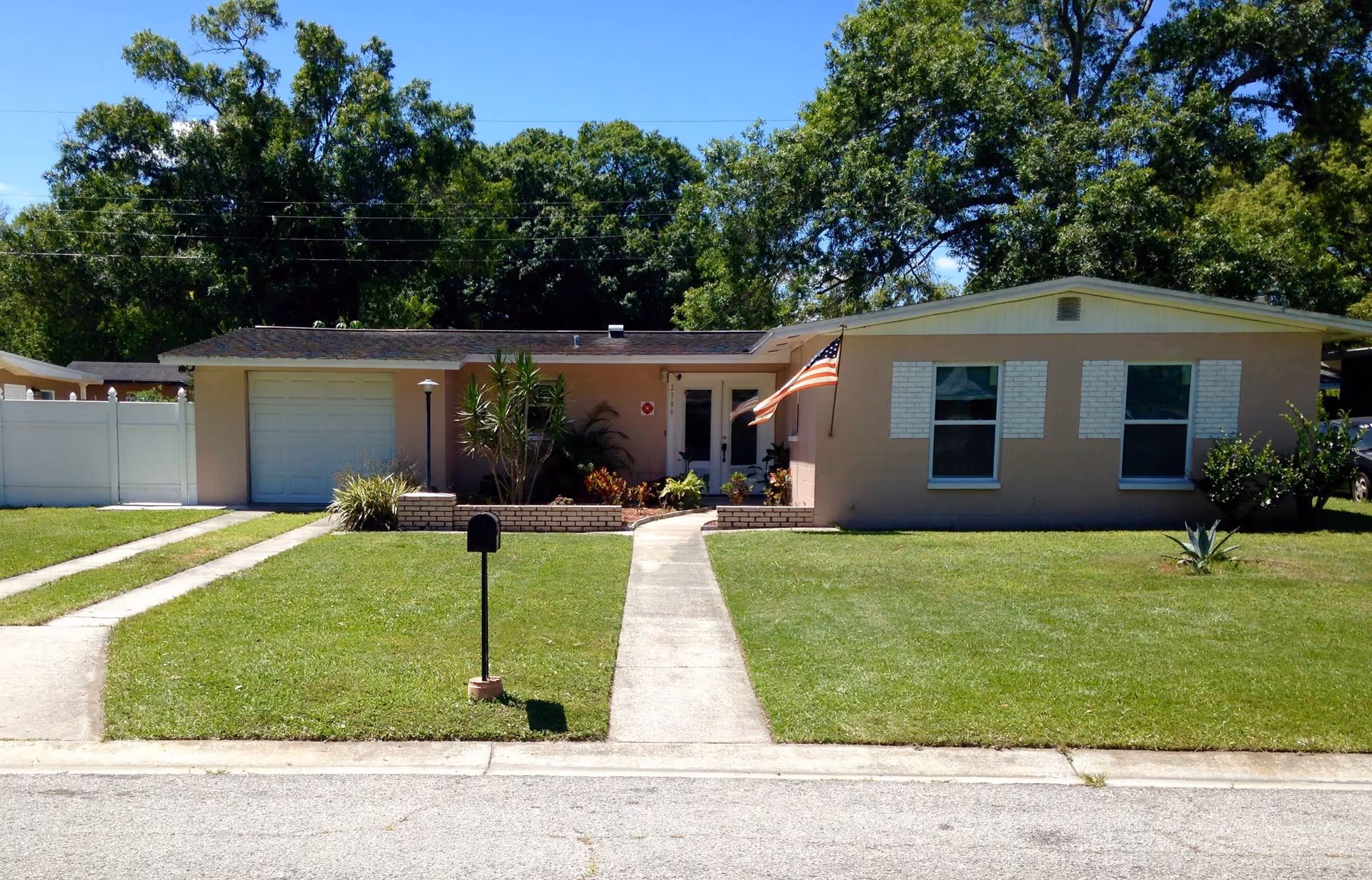 Single-story house with an American flag, front lawn, mailbox, driveway, and trees in the background.