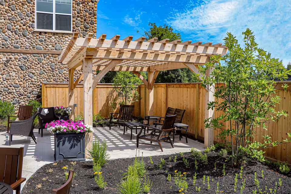 Sunny outdoor patio with a wooden pergola, lounge chairs, planters and landscaping beside a stone-faced building.