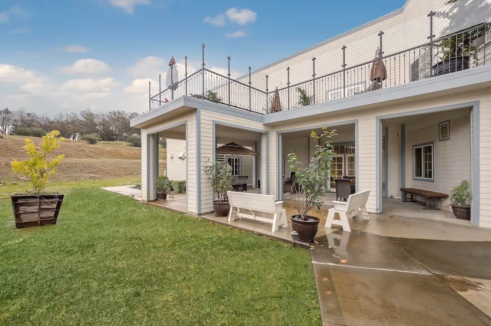 Outdoor patio area of a senior living facility with white benches, potted plants, and a covered seating area. The building is light-colored with a railing on the rooftop terrace. The surrounding area includes a grassy lawn and a small hill with trees in the background under a partly cloudy sky.