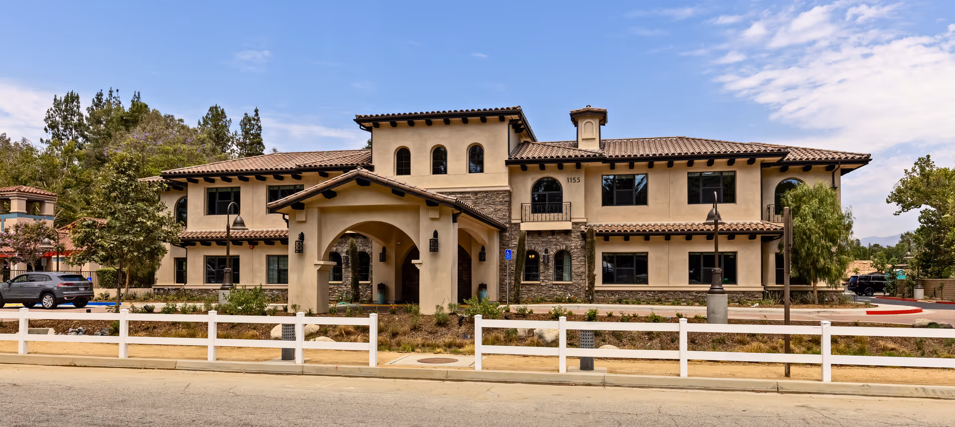 Front exterior view of a two-story senior living facility building with beige stucco walls, stone accents, and a tiled roof. The entrance features an arched covered walkway. There are trees, shrubs, and a white fence in front of the building, with a clear blue sky above.